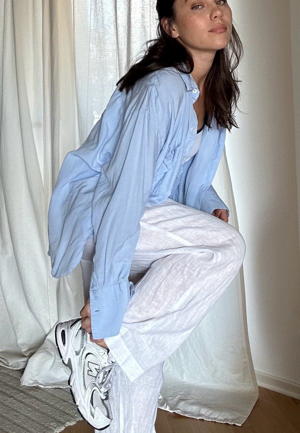 Helles blaues oversized Hemd, weiße gemusterte Hose und graue Sneaker. Weiche Stoffe, lockere Passform, sichtbare Knopfleiste und Manschetten.