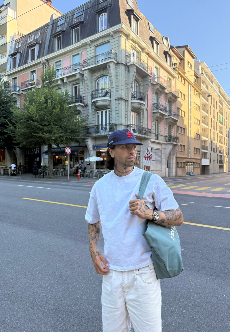 Jeune homme tatoué portant une chemise blanche et une casquette se tient dans une rue de la ville avec des bâtiments historiques et un café en arrière-plan.