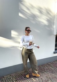 Langermet hvit topp, beigefargede vide bukser, brune støvler. Holder en iskald drikke, iført store svarte solbriller. Står mot en lys vegg.