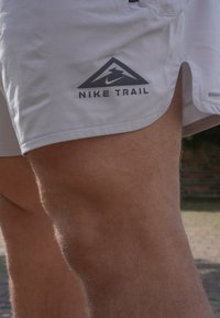 Gaiši pelēki sporta šorti ar trīsstūra melnu logotipu, uz kura rakstīts "NIKE TRAIL". Funkcijas: atvērta sānu šuvēm un gluda tekstūra.