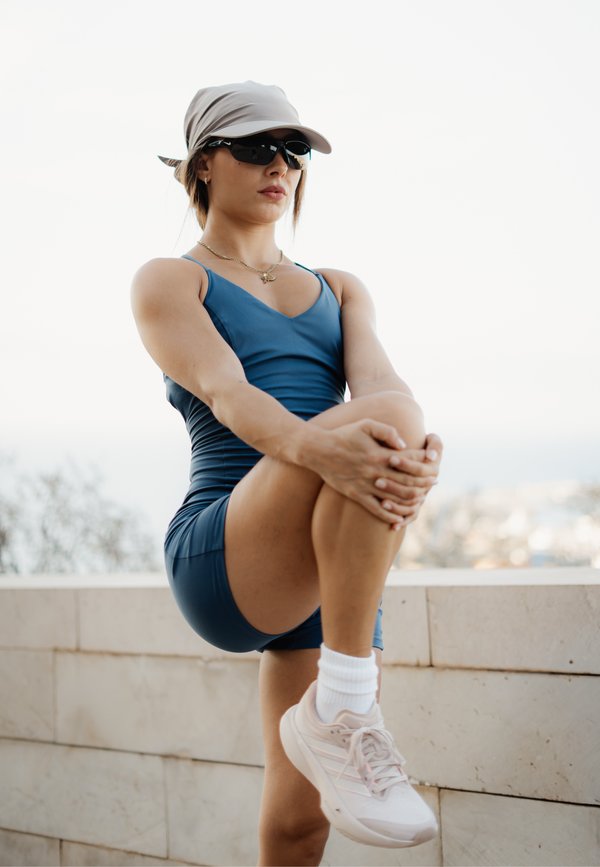 Femme en tenue de sport étirant sa jambe en plein air, portant une casquette, des lunettes de soleil et des baskets blanches.