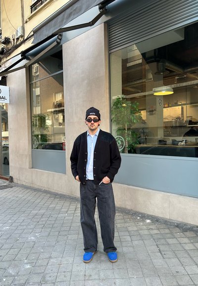Cárdigan negro de punto sobre una camisa azul clara, combinado con jeans negros holgados y zapatos azules. De pie contra un edificio con ventanas de cristal.