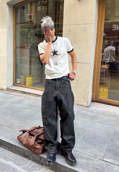 Camiseta blanca con una estrella negra, vaqueros anchos negros, zapatos negros y un gorro de lana estampado. Bolsa de cuero marrón a los pies sobre el pavimento de piedra.
