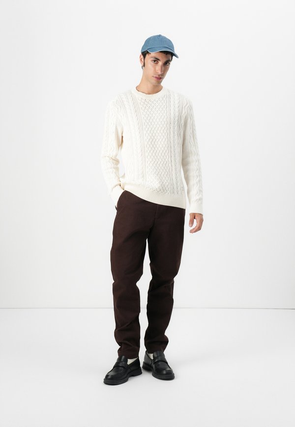 Sweater de malha em creme, calças castanhas escuras e mocassins pretos. Boné azul completa o visual. Fundo minimalista destaca a roupa.