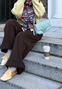 Personne assise sur des marches en pierre à l'extérieur, portant un pull jaune, une écharpe à motifs, un pantalon marron, des chaussures jaunes, avec un sac à main bleu clair et une tasse de café à proximité.