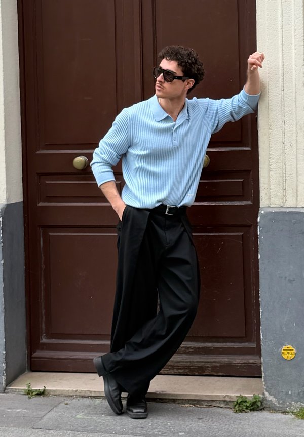 Άνδρας με μπλε ριγωτό πουλόβερ, μαύρο φαρδύ παντελόνι και γυαλιά ηλίου στηριγμένος σε ξύλινη πόρτα, όρθιος σε αστικό πεζοδρόμιο.