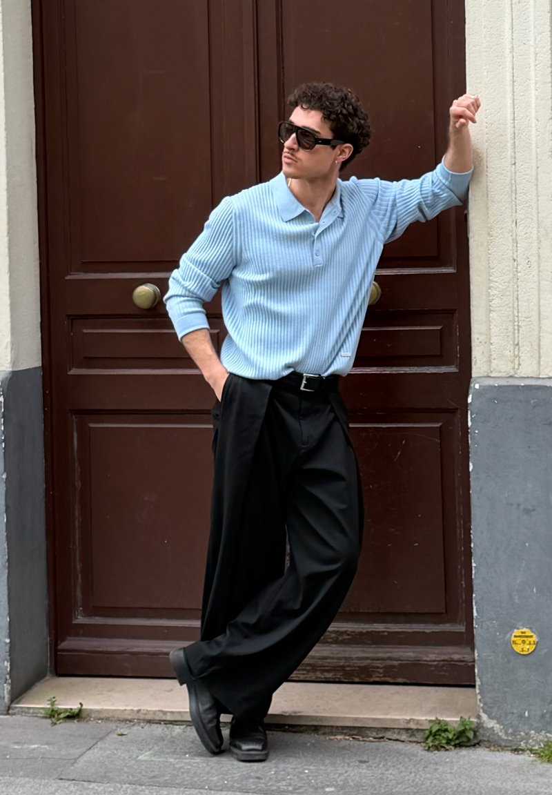 Homme en pull côtelé bleu, pantalon large noir et lunettes de soleil, appuyé contre une porte en bois, debout sur un trottoir urbain.