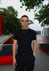 T-shirt preto justo, calças pretas, joias prateadas; cabelo de comprimento médio; sardas, em pé ao ar livre perto de almofadas com detalhes vermelhos e uma fonte de água.