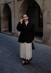 Czarna, strukturalna marynarka, kremowa spódnica z czarną koronkową lamówką, czarne, szpiczaste obcasy oraz mała czarna torebka; nosząca okulary przeciwsłoneczne, stoi na brukowanej ulicy.