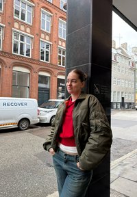 Jeune femme portant une veste verte et une chemise rouge, les mains dans les poches, appuyée contre un pilier noir dans une rue urbaine avec des bâtiments en briques et une camionnette blanche.