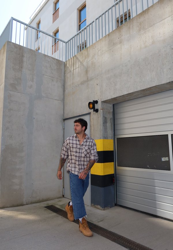 Hombre con camisa de cuadros, vaqueros azules y botas marrones caminando junto a una pared de hormigón con rayas negras y amarillas cerca de una puerta de garaje cerrada.