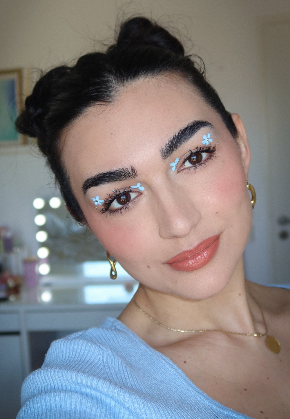 Portret van een persoon met donker haar in twee knotjes, lichte blauwe bloem make-up onder de ogen, gekleed in een lichtblauw topje, met gouden oorbellen en een ketting.