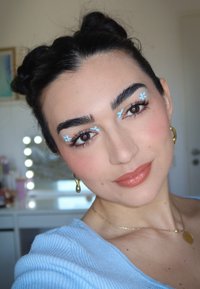 Portret van een persoon met donker haar in twee knotjes, lichte blauwe bloem make-up onder de ogen, gekleed in een lichtblauw topje, met gouden oorbellen en een ketting.
