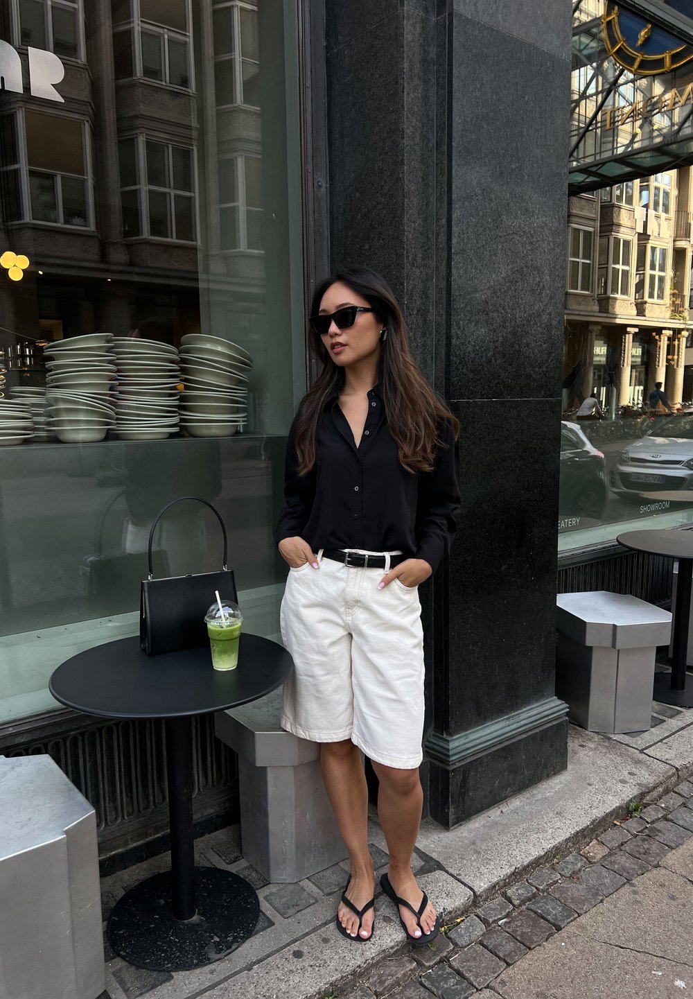 Μαύρο πουκάμισο με μακριά μανίκια και κουμπιά, άσπρο σορτς μέχρι το γόνατο, μαύρη ζώνη, μαύρη τσάντα και σανδάλια. Πράσινο ποτό σε στρογγυλό μαύρο τραπέζι.
