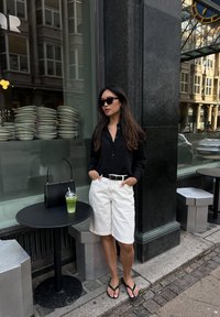 Μαύρο πουκάμισο με μακριά μανίκια και κουμπιά, άσπρο σορτς μέχρι το γόνατο, μαύρη ζώνη, μαύρη τσάντα και σανδάλια. Πράσινο ποτό σε στρογγυλό μαύρο τραπέζι.