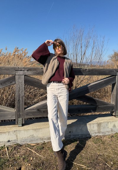 Mujer con gafas de sol, suéter color granate, chaleco beige y pantalones blancos, de pie al aire libre junto a una cerca de madera con juncos secos y cielo azul claro detrás.