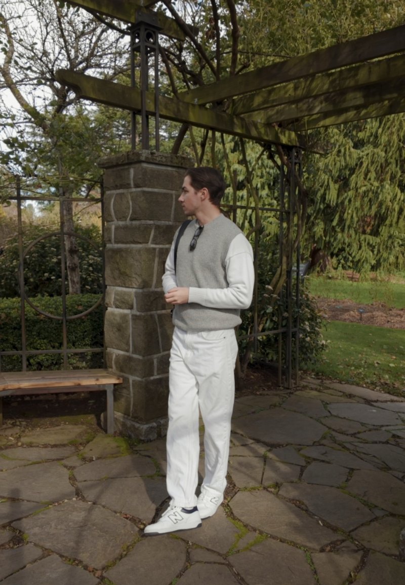 Mann in weißer Hose und grauem Pullover steht auf einer Stein-Terrasse unter einem hölzernen Pergola im Garten und blickt nach links.