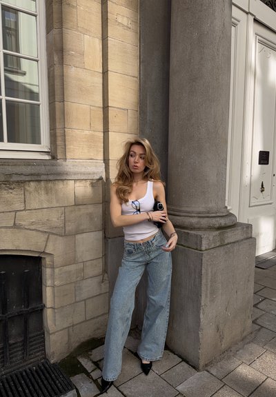 Joven con cabello largo, camiseta blanca sin mangas y vaqueros azules de pierna ancha, de pie frente a un edificio de piedra con una gran columna y ventana.