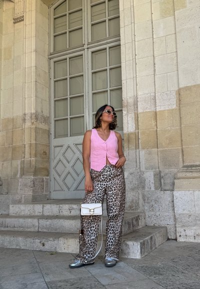 Chaleco de lino rosa, pantalones con estampado animal, bolso blanco, zapatillas plateadas; de pie en unos escalones cerca de una pared de piedra texturizada y ventanas.