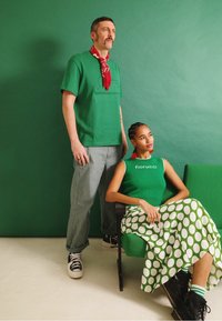 Camisa verde de manga curta com um design texturizado, calças cinzentas, lenço vermelho e saia com bolinhas em vários tons de verde. Ténis pretos.