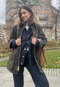 Oliwkowozielona woskowana kurtka na jeansowej kurtce, w zestawie z ciemnymi dżinsowymi spodniami. Posiada złote okucia, kieszenie i fakturowaną tkaninę.