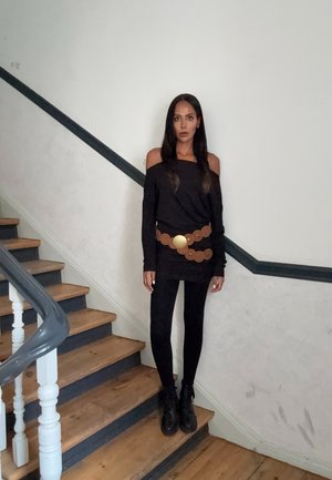 Top negro sin hombros combinado con un cinturón marrón de patrón circular y leggings negros. Botas negras completan el outfit frente a una escalera de madera.