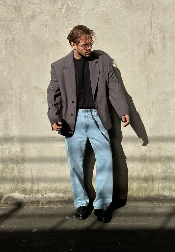 Blazer cinza oversize com riscas, t-shirt preta, jeans de perna larga em azul claro e sapatos pretos em frente a uma parede cinza texturizada.