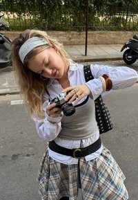 Ezüst testű és fekete objektívű fényképezőgép, amelyet egy rétegesen öltözött személy tart: szürke trikó, fehér csíkos ing és kockás szoknya.