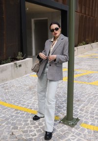 Hnědošedý blazer s houndstooth vzorem přes béžovou košili, světle modré zvonové džíny a černé boty. Doplňky zahrnují černé sluneční brýle a malou tašku.