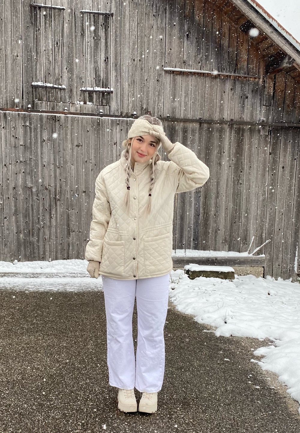Giacca trapuntata color crema con tasche frontali, abbinata a pantaloni bianchi e stivali chiari. Fiocchi di neve che cadono in un ambiente rustico all'aperto.