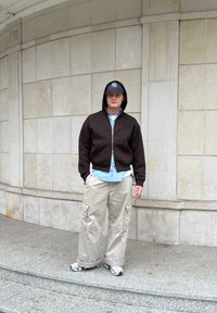 Man wearing a black hoodie, blue shirt, beige cargo pants, sneakers, glasses, and a navy cap stands on stone steps against a tiled wall.