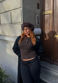 Brun ribbad stickad topp med polokrage, svarta byxor och en lurvig brun hatt. Står mot en texturerad stenvägg nära en träport.