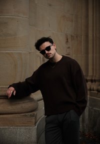 Donkerbruin oversized trui van gebreid materiaal, gecombineerd met grijze broek. Accessoires zijn onder meer een zonnebril en een kleine hangerketting.