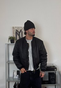 Fekete bomberdzseki cipzárral az elején és bordázott mandzsettával, egy szürke póló felett viselve. Fekete sapka, minimalistának mondható beltéri környezetben állva.