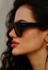 Μαύρα γυαλιά ηλίου Ray-Ban με υπερμεγέθη τετράγωνα σκελετά, σκουρόχρωμους φακούς και ασημένιο λογότυπο στον κρόταφο, συμπληρωμένα από σγουρά μαλλιά και λεπτές δερματικές λεπτομέρειες.
