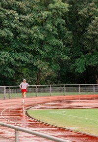 Biegacz w czerwonych szortach i białej czapce biega po mokrej ścieżce na świeżym powietrzu, otoczonej zielonymi drzewami i metalowymi poręczami.