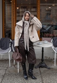 Veste en fausse fourrure crème avec bordure noire contrastante, pantalon large en cuir marron, écharpe grise tricotée et bottes noires à talons aiguilles. Sac à main marron.