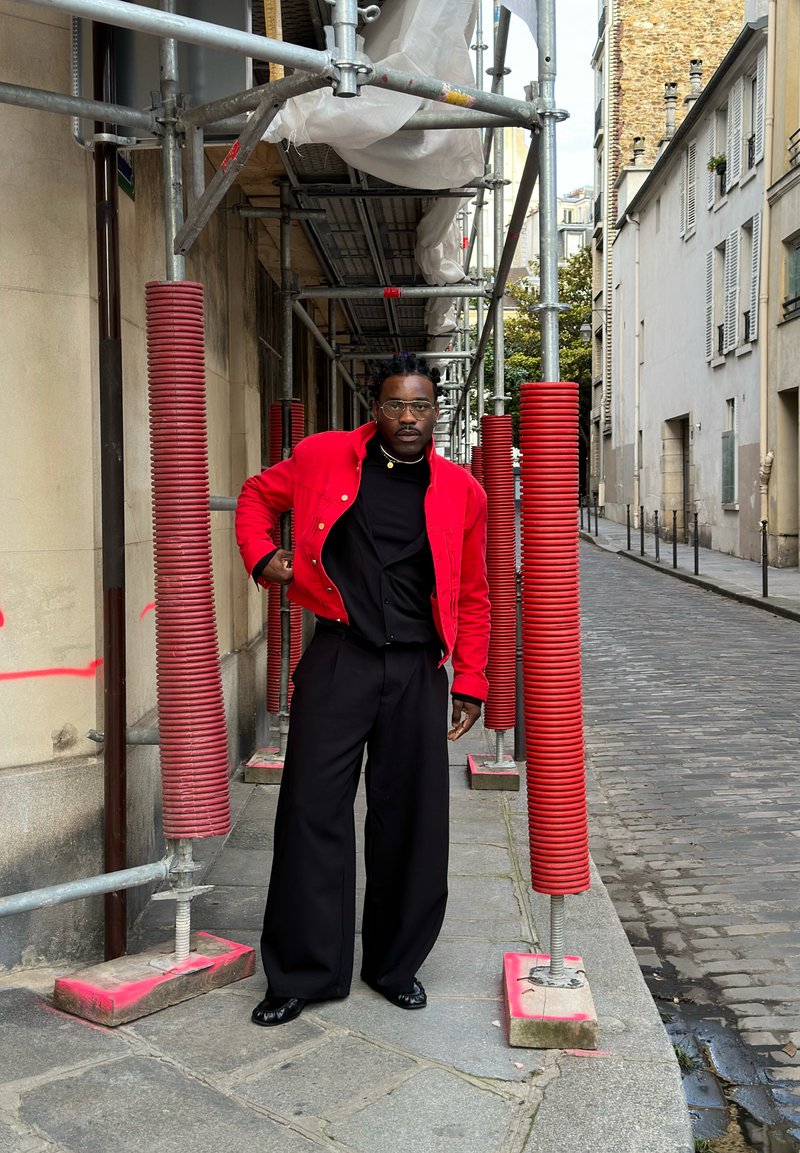 Άτομο με γυαλιά που φοράει κόκκινο μπουφάν και μαύρο παντελόνι με φαρδιά μπατζάκια στέκεται κάτω από σκαλωσιά με κόκκινη επένδυση σε στενό λιθόστρωτο δρόμο.