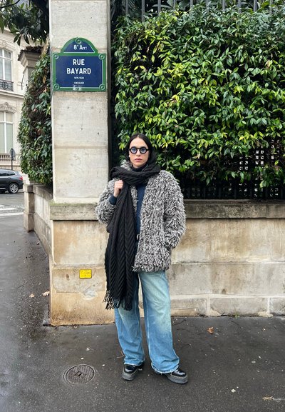Abrigo gris de gran tamaño y borroso, bufanda negra, suéter azul, jeans claros de pierna ancha y zapatos de plataforma negros, de pie al lado de una señal de tráfico con borde verde.