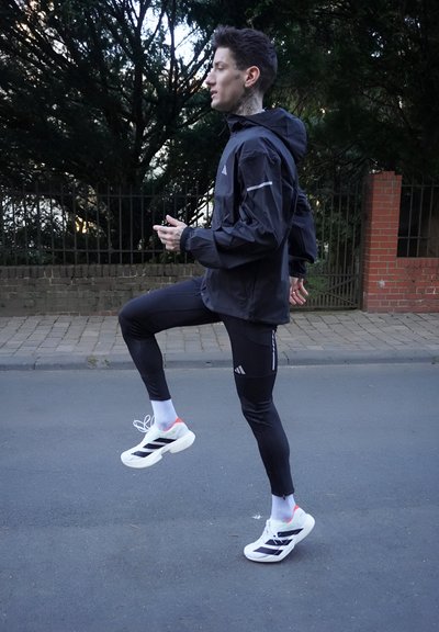 Chaqueta ligera negra con capucha, leggins ajustados negros y zapatillas blancas con rayas negras y acentos neón. Acera y follaje verde de fondo.