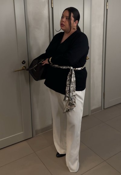 Mujer con blusa negra y pantalones blancos con cinturón estampado, sosteniendo una bolsa oscura cerca de unas puertas blancas cerradas en una habitación con azulejos.