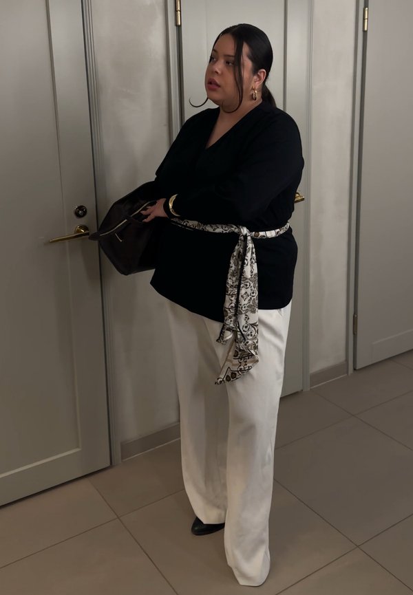 Femme portant un haut noir et un pantalon blanc avec une ceinture à motifs, tenant un sac sombre près de portes blanches fermées dans une pièce carrelée.