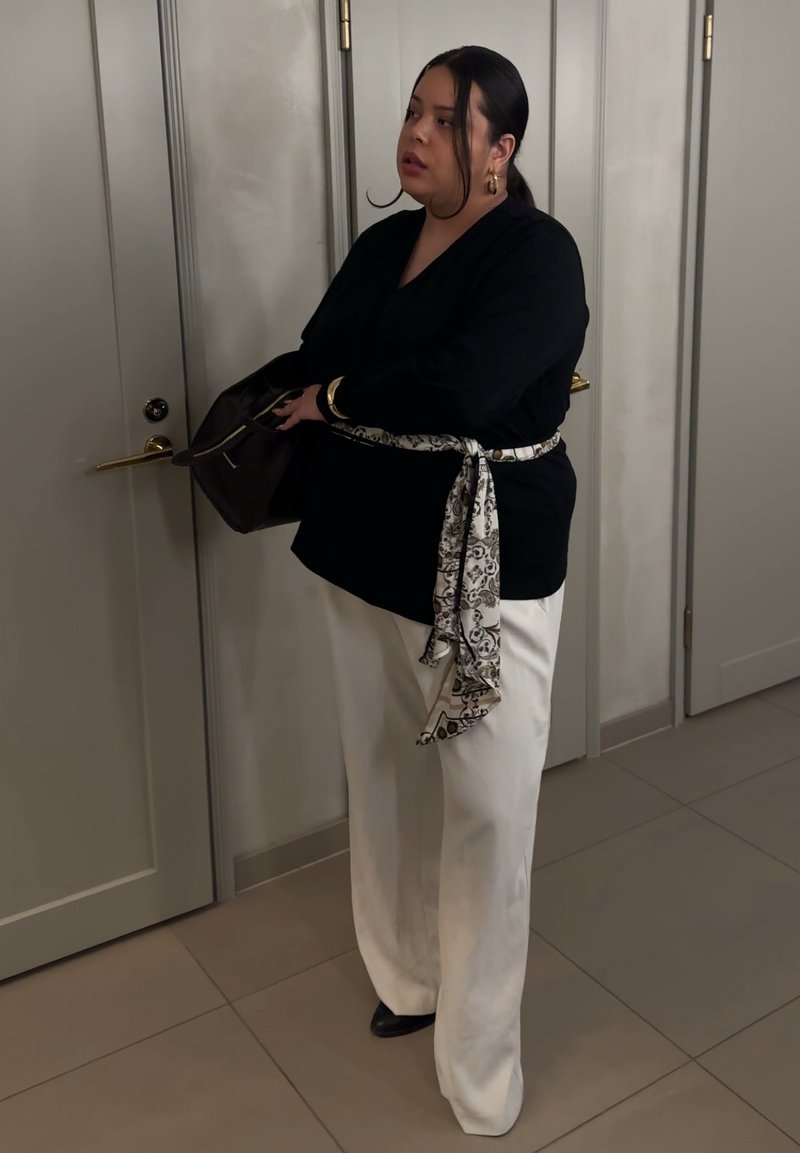Femme portant un haut noir et un pantalon blanc avec une ceinture à motifs, tenant un sac sombre près de portes blanches fermées dans une pièce carrelée.