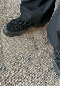 Svarte semskede joggesko med teksturert finish, med tykke svarte gummisåler og kryssende snøring. Brukt med svarte bukser på et mønstret underlag.