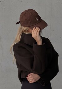 Brun bomull baseballkeps med en svängd skärm, prydd med en broderad logga. Bärs med en svart oversized kappa och stylat hår.