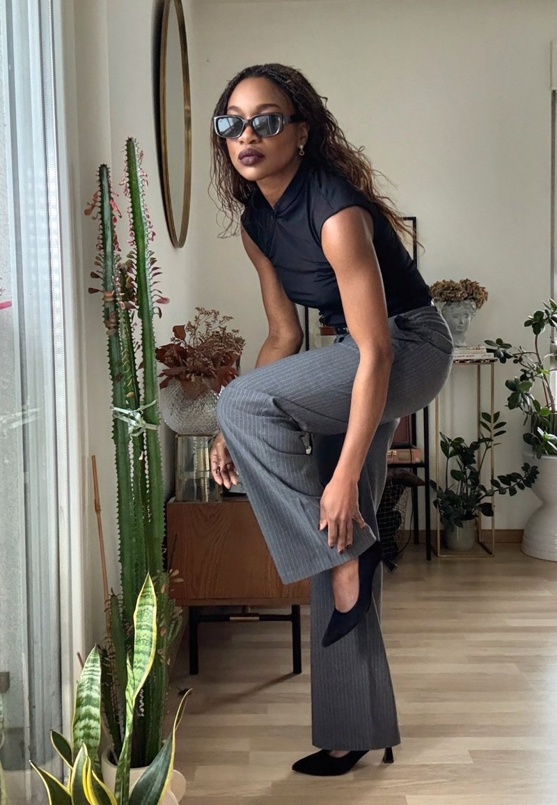 Femme portant des lunettes de soleil ajustant une chaussure noire à talon haut, vêtue d'un haut noir sans manches et d'un pantalon gris à fines rayures, debout près de plantes à l'intérieur.