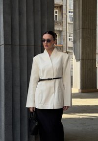 Witte op maat gemaakte blazer met brede mouwen en een ceintuur op de taille, gecombineerd met een zwarte rok. Accessoires zijn onder andere zwarte zonnebril en een zwarte handtas.