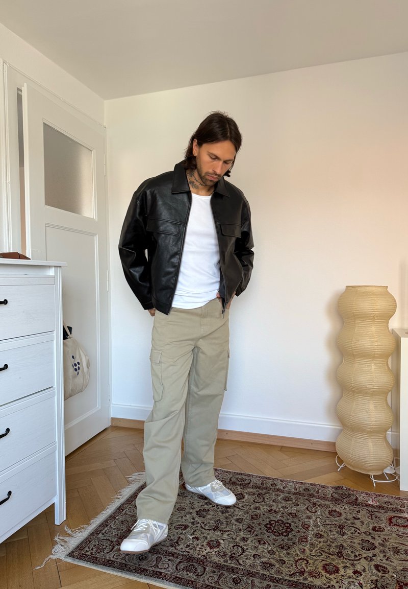 Homme aux cheveux foncés portant une veste en cuir noire, une chemise blanche, un pantalon cargo beige et des baskets blanches, debout sur un tapis à motifs dans une pièce.