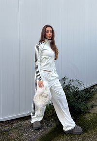 Γυναίκα σε λευκή φόρμα με μαύρες ρίγες κρατάει λευκό χνουδωτό καπέλο, στέκεται σε εξωτερικό χώρο μπροστά σε γκρι επενδυμένο τοίχο και πράσινη βλάστηση.