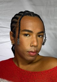 Personne aux cheveux tressés ornés de perles transparentes, portant des boucles d'oreilles en anneau doré et un anneau au nez, vêtue d'un haut en maille rouge sur un fond uni.